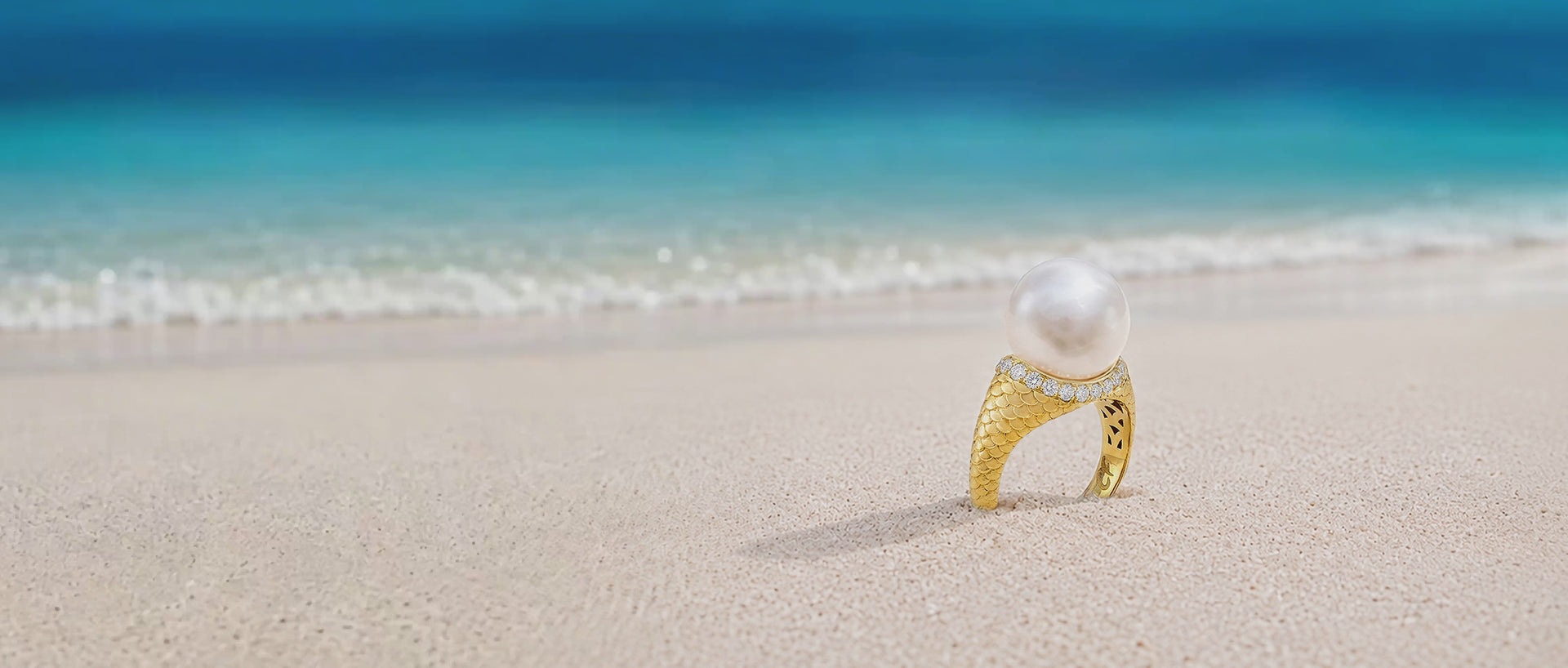 Colette Falchet Alma Mater ring in 18k yellow gold with a white South Sea pearl and natural diamonds, standing on sand beside the turquoise sea.